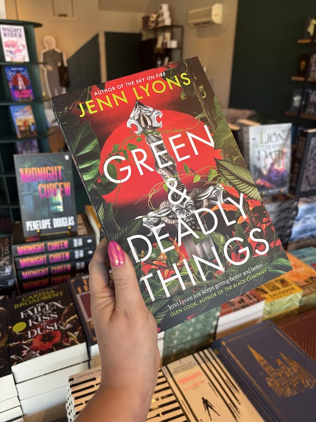 A hand with pink nail polish holding a copy of 'Green & Deadly Things' by Jenn Lyons, featuring a large silver dagger with a ram's head handle, set against a red sun and tropical green leaves.