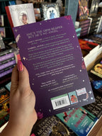 A hand with pink nail polish holding the back of a copy of 'Grim Tidings' by B.K. Borison, featuring a dark purple background, white text, and a scattering of sparkles, pink flowers, and white feathers.