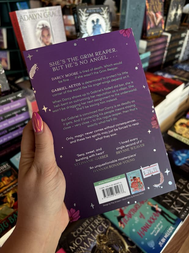 A hand with pink nail polish holding the back of a copy of 'Grim Tidings' by B.K. Borison, featuring a dark purple background, white text, and a scattering of sparkles, pink flowers, and white feathers.