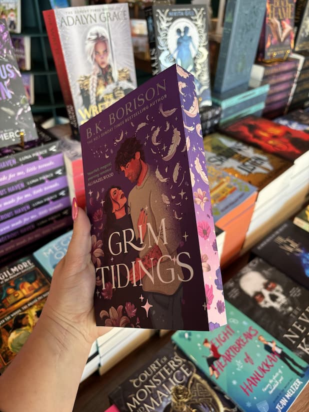 A hand with pink nail polish holding a copy of 'Grim Tidings' by B.K. Borison, featuring an illustrated cover of a couple embracing and looking at each other. They are surrounded by feathers and flowers on a purple background with matching sprayed edges. 