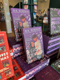 A stack of 'Grim Tidings' by B.K. Borison, featuring an illustrated cover of a couple embracing and looking at each other. They are surrounded by feathers and flowers on a purple background with matching sprayed edges. 