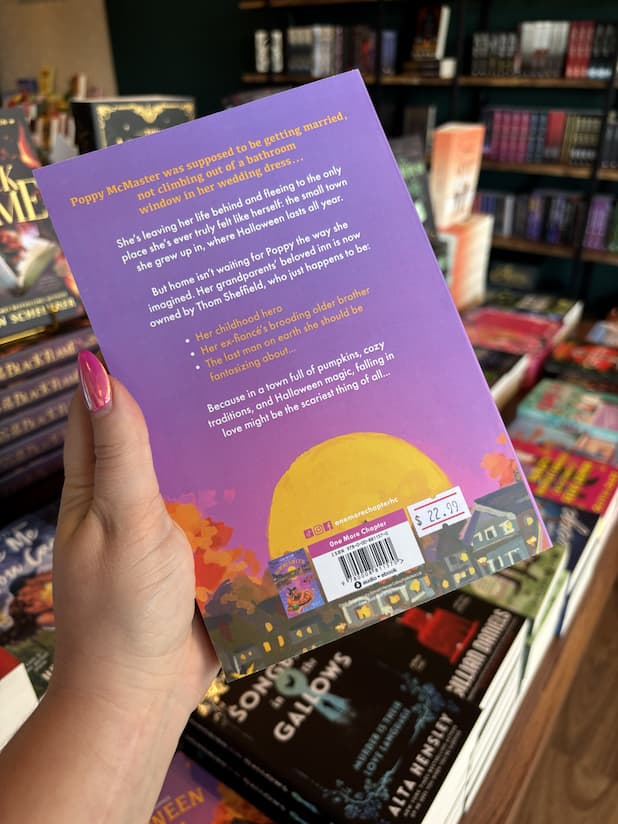 A hand with pink nail polish holding the back of a copy of 'Halloween Town' by Joss Wood with a purple sunset and white and yellow text.