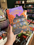 A hand with pink nail polish holding a copy of 'Halloween Town' by Joss Wood with a Halloween-themed house and decorations.