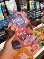 A hand holds the book cover of 'Hate You to Love You' by Joshua Hortinela with illustration of two men lying down and digitally sprayed edges featuring open books and headphones.