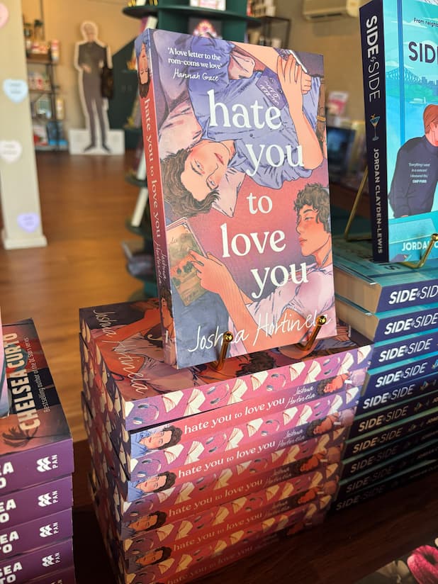 A stack of 'Hate You to Love You' by Joshua Hortinela books with alternating spine and sprayed edges facing out. The cover features an illustration of two men lying down and digitally sprayed edges featuring open books and headphones.
