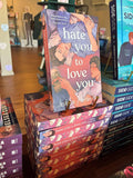 A stack of 'Hate You to Love You' by Joshua Hortinela books with alternating spine and sprayed edges facing out. The cover features an illustration of two men lying down and digitally sprayed edges featuring open books and headphones.