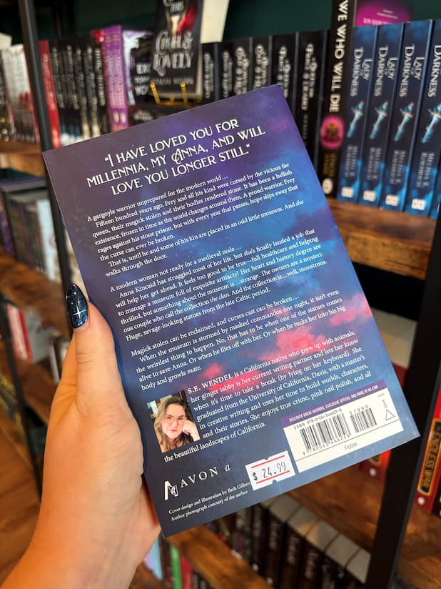 A hand holds the back cover of 'Heartsong' featuring white text for the blurb against a blue and pink cloudy sky. The book is held in front of a bookshelf full of other books blurry in the background. 
