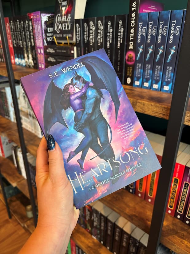 A hand holds a book cover of 'Heartsong' featuring a gargoyle and a woman embracing against a colourful cloudy sky. The book is held in front of a bookshelf full of other books blurry in the background 