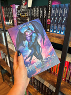 A hand holds a book cover of 'Heartsong' featuring a gargoyle and a woman embracing against a colourful cloudy sky. The book is held in front of a bookshelf full of other books blurry in the background 