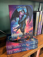 A stack of the book 'Heartsong' with a single copy sitting on a gold stand with cover displayed. The cover features a gargoyle and a woman embracing against a colourful cloudy sky. The book is sitting on a wooden shelf in front of a green wall. 