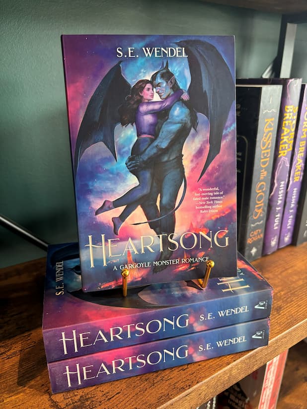 A stack of the book 'Heartsong' with a single copy sitting on a gold stand with cover displayed. The cover features a gargoyle and a woman embracing against a colourful cloudy sky. The book is sitting on a wooden shelf in front of a green wall. 