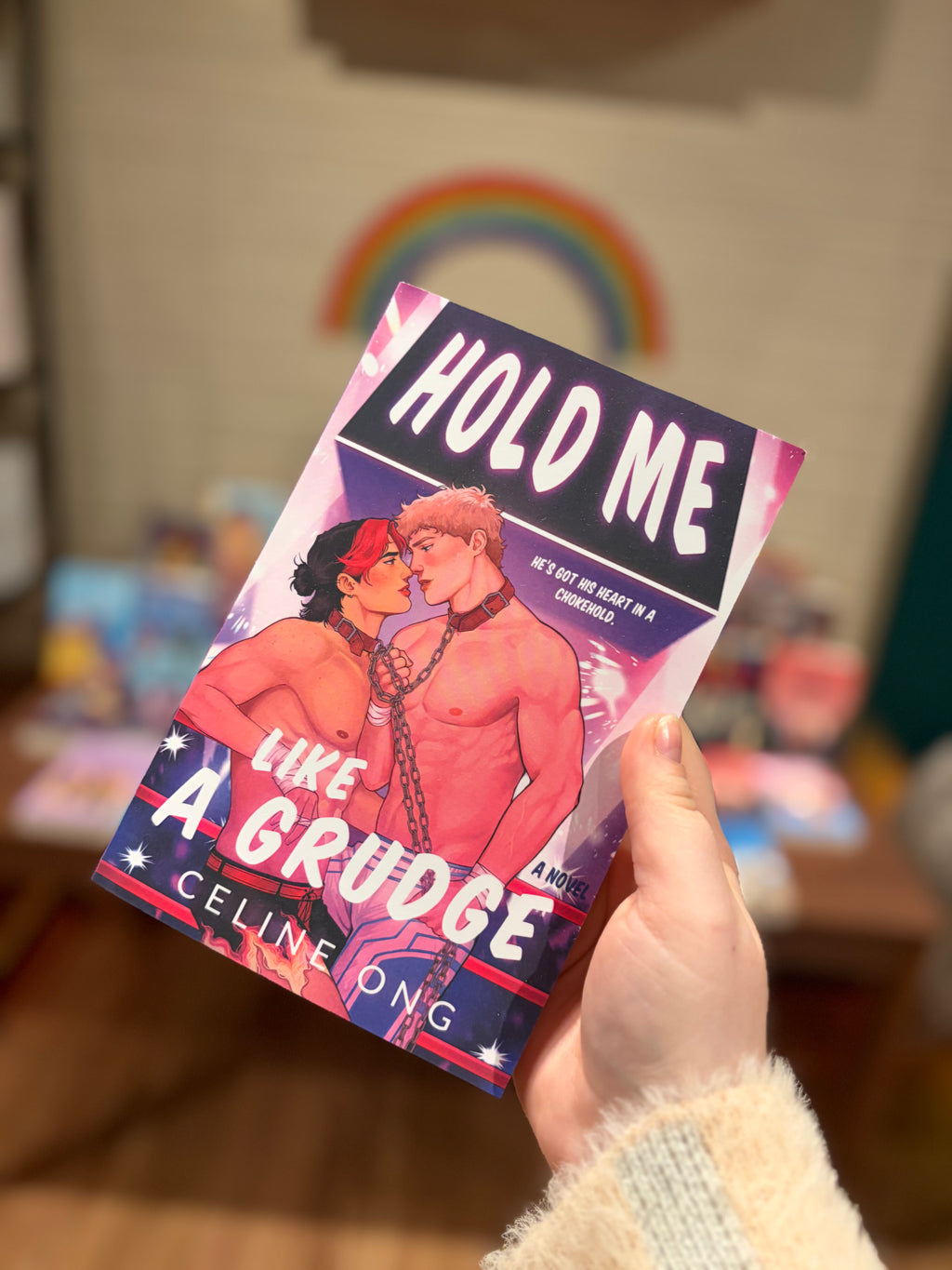 A hand holding a copy of the book 'Hold Me Like A Grudge' by Celine Ong, showing the front cover illustration of two male wrestlers bound by a chain around the neck against a blurred bookstore background.