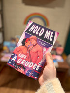 A hand holding a copy of the book 'Hold Me Like A Grudge' by Celine Ong, showing the front cover illustration of two male wrestlers bound by a chain around the neck against a blurred bookstore background.