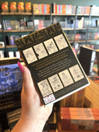 A hand with pink nail polish holding the back of Holly Black's Faerie Coloring Postcards box set, featuring a black and gold design with line-art illustrations of fairies and fantasy scenes on the front and spine.