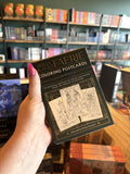 A hand with pink nail polish holding Holly Black's Faerie Coloring Postcards box set, featuring a black and gold design with line-art illustrations of fairies and fantasy scenes on the front and spine.