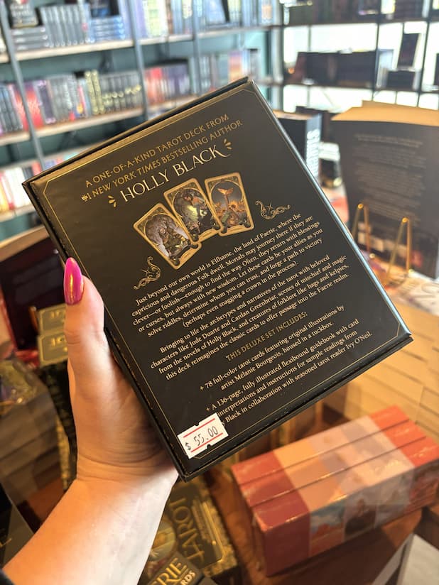 A hand with pink nail polish holding the back of Holly Black's Faerie Tarot Deck and Guidebook box set, featuring a black box with ornate gold patterns and central artwork of a cauldron with white faeries  and two circular character illustrations.