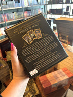 A hand with pink nail polish holding the back of Holly Black's Faerie Tarot Deck and Guidebook box set, featuring a black box with ornate gold patterns and central artwork of a cauldron with white faeries  and two circular character illustrations.