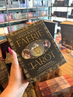A hand with pink nail polish holding Holly Black's Faerie Tarot Deck and Guidebook box set, featuring a black box with ornate gold patterns and central artwork of a cauldron with white faeries  and two circular character illustrations.