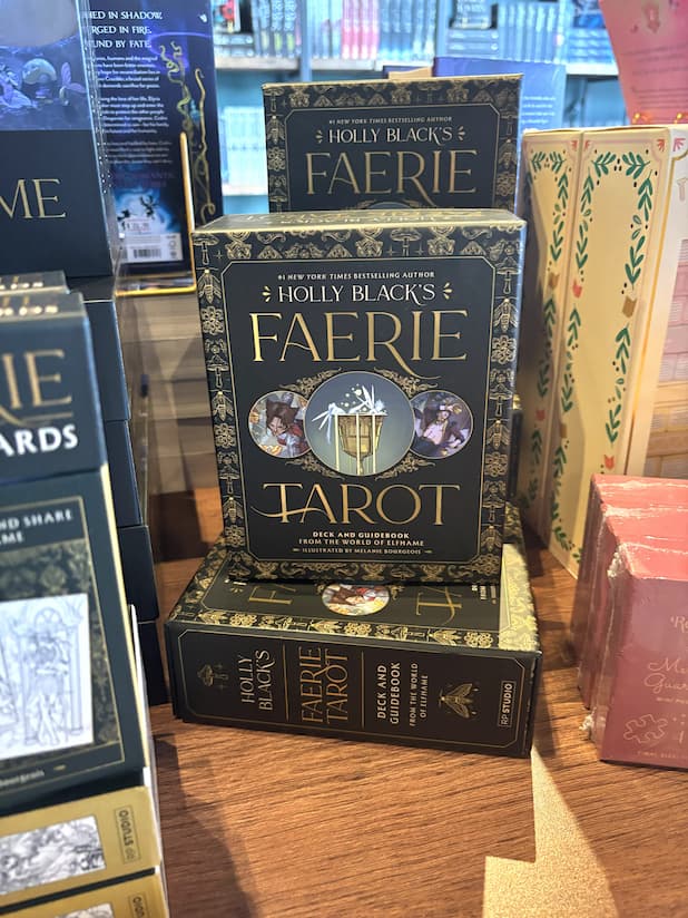 A stack of Holly Black's Faerie Tarot Deck and Guidebook box set, featuring a black box with ornate gold patterns and central artwork of a cauldron with white faeries  and two circular character illustrations.