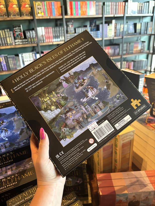 A hand with pink nail polish holds the back of an 1000 piece puzzle box featuring an illustration of a fantasy landscape with buildings and people, titled 'Holly Black's Isles of Elfhame'.