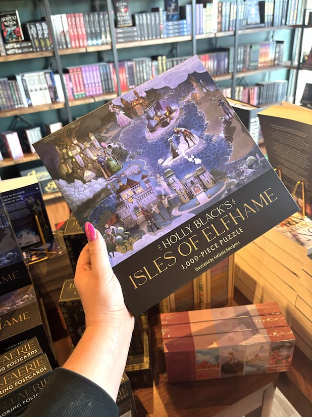A hand with pink nail polish holds up an 1000 piece puzzle box featuring an illustration of a fantasy landscape with buildings and people, titled 'Holly Black's Isles of Elfhame'.