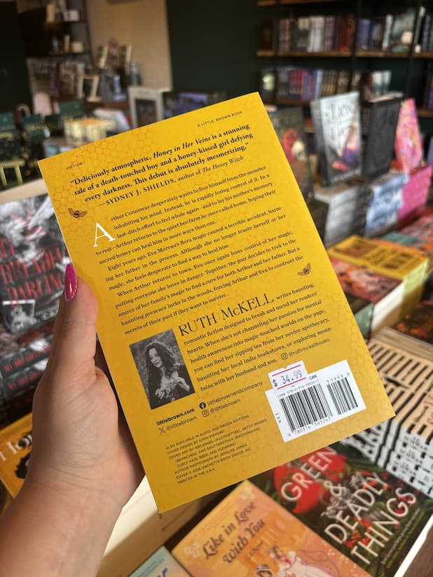 A hand with pink nail polish holds the back of a copy of 'Honey in Her Veins' by Ruth McKell, featuring a yellow background, bees, and black text.