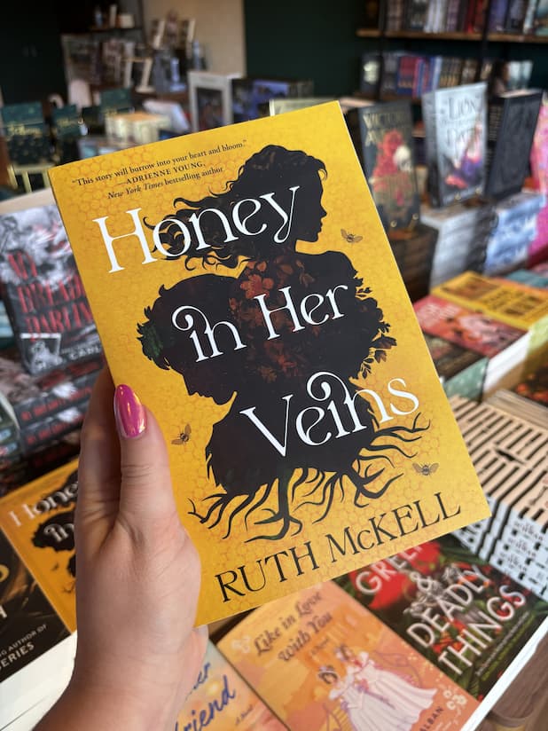 A hand with pink nail polish holds a copy of 'Honey in Her Veins' by Ruth McKell, featuring a silhouette of a woman and a man, bees around them on a yellow background.
