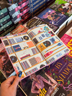 A hand holds open the special edition book 'Hook, Line, and Sinker' by Tessa Bailey, showing the end papers illustrated with records, musical instruments and beach paraphernalia. The book is held in front of a table full of other books blurry in the background.
