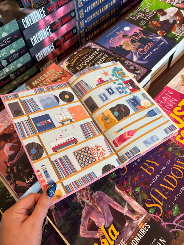 A hand holds open the special edition book 'Hook, Line, and Sinker' by Tessa Bailey, showing the end papers illustrated with records, musical instruments and beach paraphernalia. The book is held in front of a table full of other books blurry in the background.