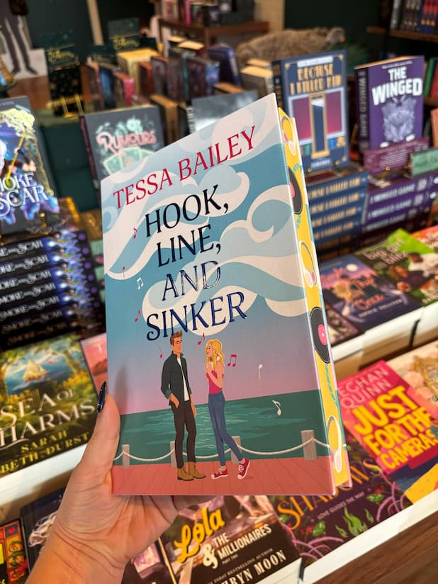 A hand holds the book cover of 'Hook, Line, and Sinker' by Tessa Bailey with a man facing a woman wearing white headphones on a dock. Pink and white music notes run up the book cover and across the title. The sprayed edges are yellow with black records and white headphones. The book is held in front of a table full of other books blurry in the background.