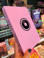 A hand holds the naked book cover of 'Hook, Line, and Sinker' by Tessa Bailey with pink cover and a black record in the centre. The book is held in front of a table full of other books blurry in the background.