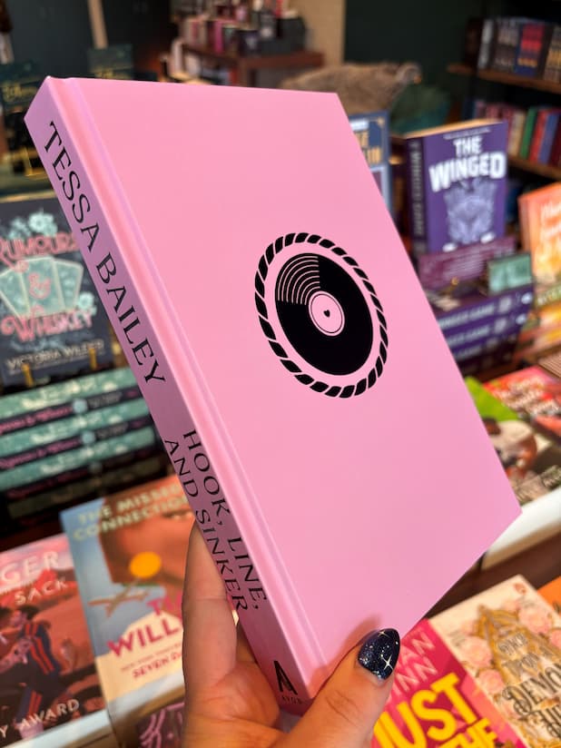 A hand holds the naked book cover of 'Hook, Line, and Sinker' by Tessa Bailey with pink cover and a black record in the centre. The book is held in front of a table full of other books blurry in the background.