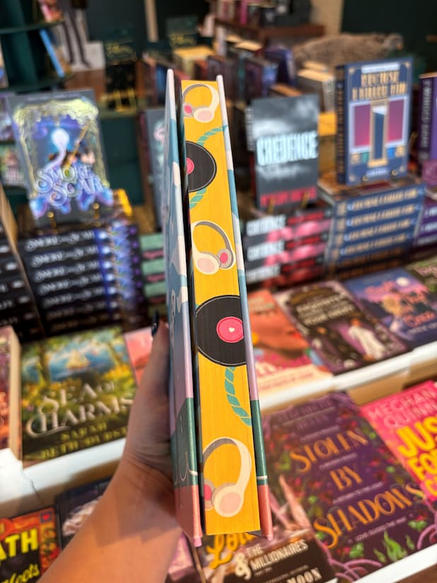 A hand holds the book cover of 'Hook, Line, and Sinker' by Tessa Bailey showing the sprayed edges, they are yellow with black records and white headphones. The book is held in front of a table full of other books blurry in the background.