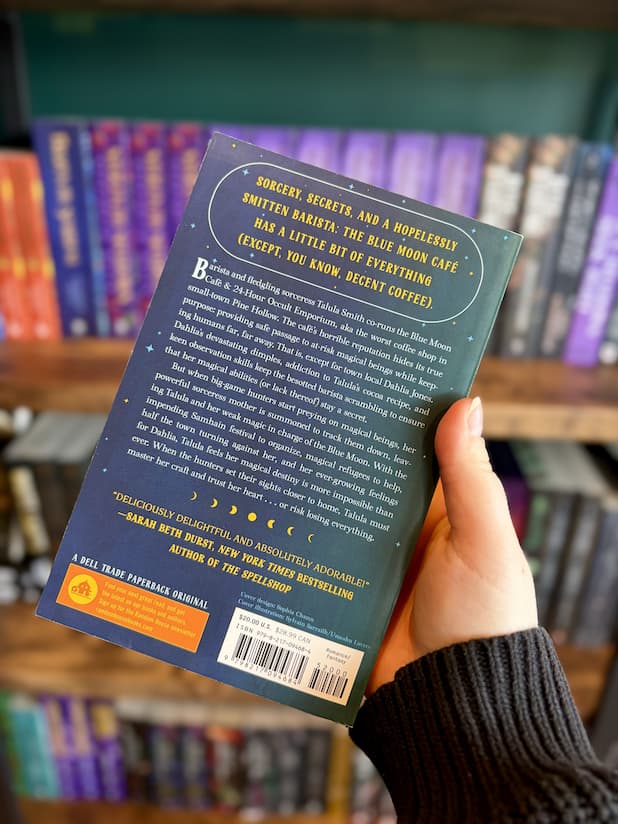 A hand holding the back of a copy of 'The Blue Moon Cafe & 24-Hour Occult Emporium' by Lexie Sharabianlou, featuring a blue starry background with white and yellow text.