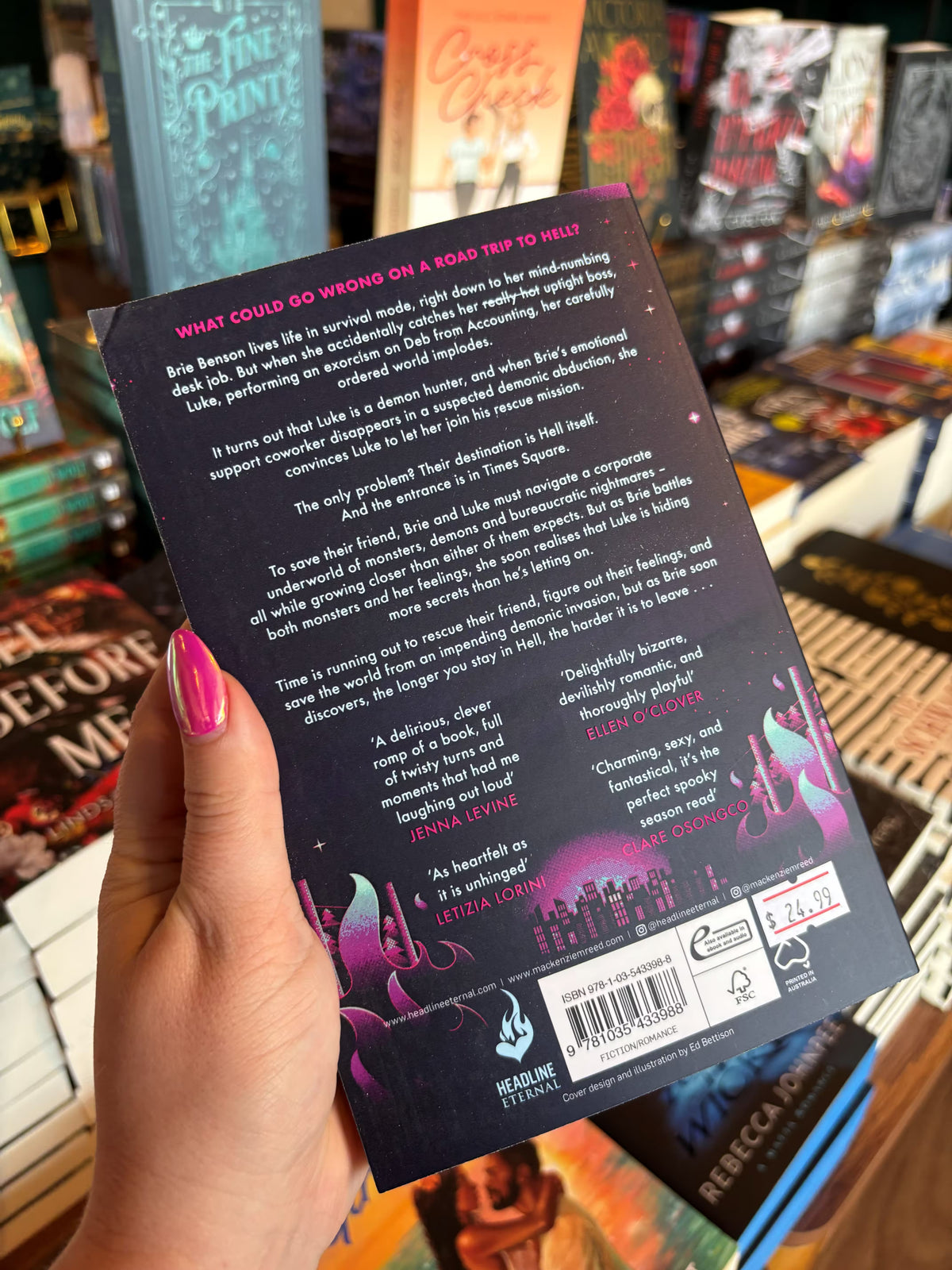 A hand with pink nail polish holds the back cover for 'I Hope This Email Finds You in Hell' by Mackenzie Reed in a bookstore with other books blurry in the background. The cover features a graphic design of city landscape in the background surrounded by flames and white text for the blurb.