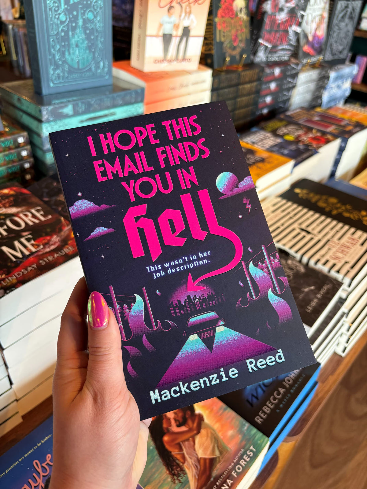 A hand with pink nail polish holds the book cover for 'I Hope This Email Finds You in Hell' by Mackenzie Reed in a bookstore with other books blurry in the background. The cover features a greaphic design of city landscape in the background surrounded by flames and a car driving into the foreground.