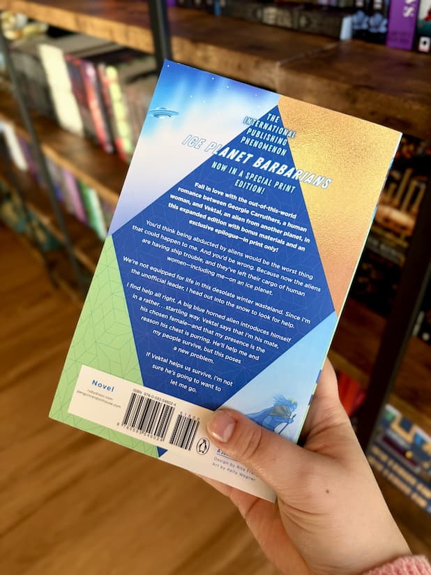 A hand holding the back of a copy of 'Ice Planet Barbarians' by Ruby Dixon, featuring a colourful triangles and a large blue diamond in the centre with white text.
