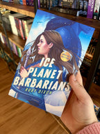 A hand holding a copy of 'Ice Planet Barbarians' by Ruby Dixon, feautring an illustration of a woman woman holding a spear, and a tall blue figure behind her, in an icy landscape.