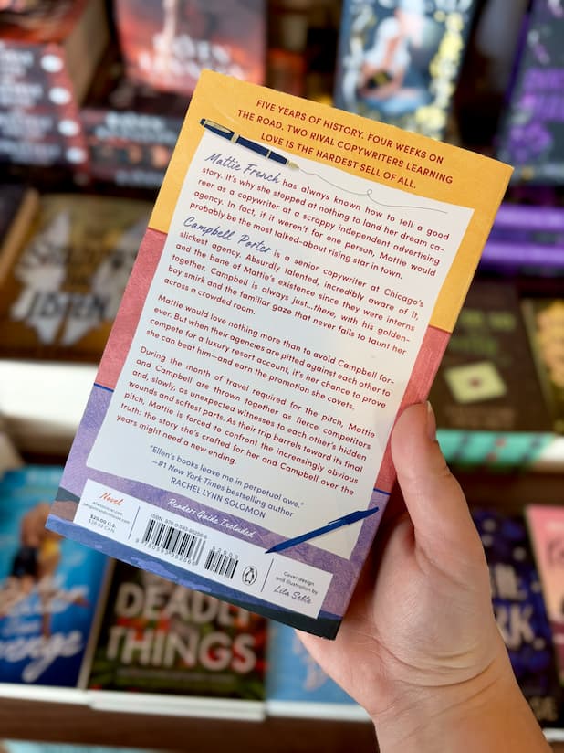 A hand holding the back of a copy of 'If Not You' by Ellen O'Clover, featuring three a yellow, red, and blue background with a piece of paper with red and blue writing.