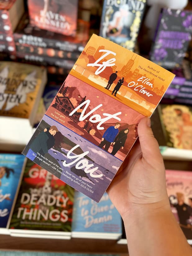 A hand holding a copy of 'If Not You' by Ellen O'Clover, featuring three illustrations of a couple in three different settings. It is split into yellow at the top, red in the middle, and blue at the bottom, with white text split between them.