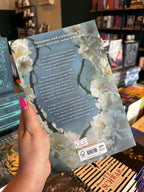 A hand with pink nail polish holds the backk cover of 'Immortal Rose' by Alexandra Bracken in a bookshop with other book titles blurry in the background. The cover has a light blue background with white flowers and green thorns on either side white text for the blurb.
