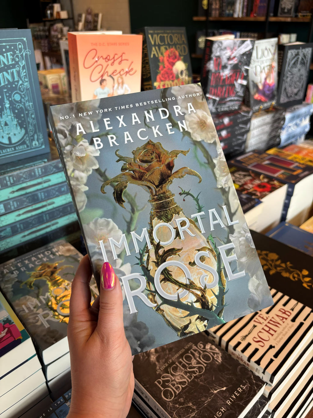 A hand with pink nail polish holds the book cover of 'Immortal Rose' by Alexandra Bracken in a bookshop with other book titles blurry in the background. The cover has a light blue background with white flowers and green thorns on either side of a gold perfume bottle with a gilded rose topper.