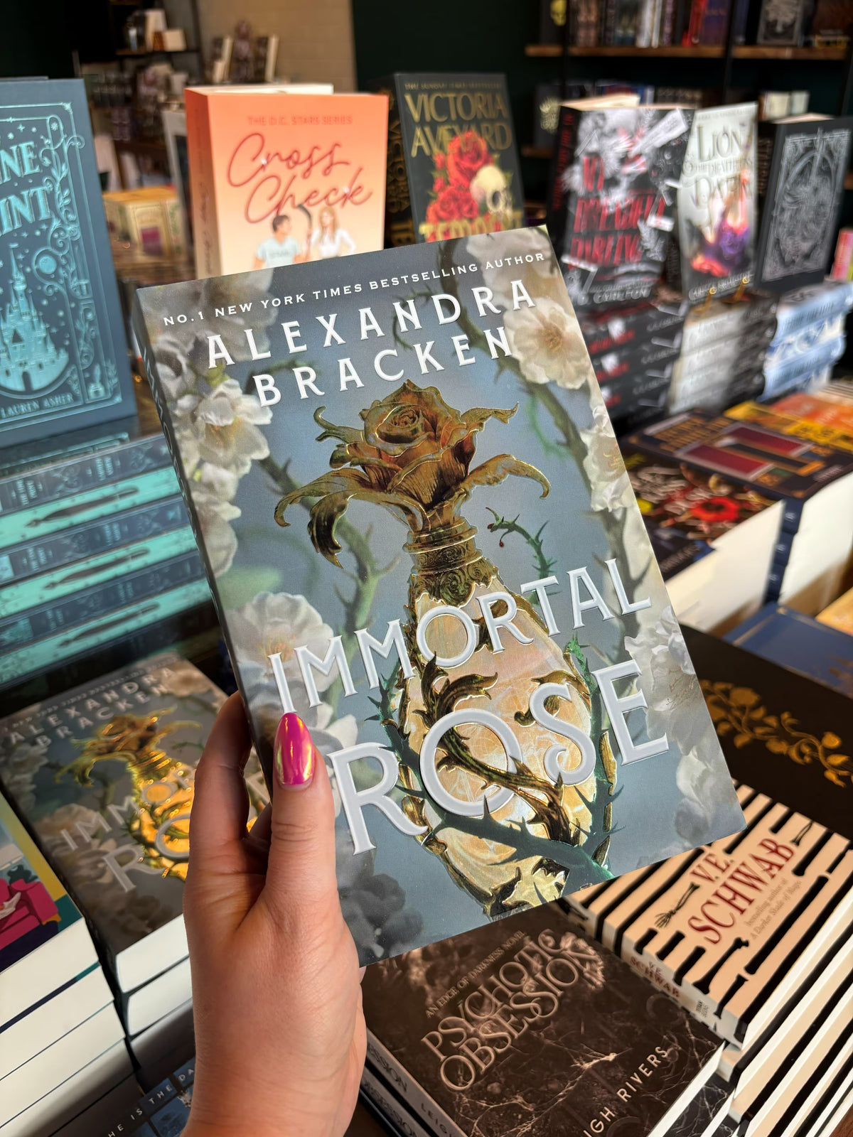 A hand with pink nail polish holds the book cover of 'Immortal Rose' by Alexandra Bracken in a bookshop with other book titles blurry in the background. The cover has a light blue background with white flowers and green thorns on either side of a gold perfume bottle with a gilded rose topper.