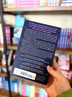 A hand holding the back of a paperback copy of 'In What World' by Bridget Morrissey. The cover is dark blue with white, yellow, and pink text.