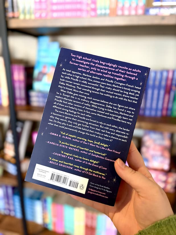 A hand holding the back of a paperback copy of 'In What World' by Bridget Morrissey. The cover is dark blue with white, yellow, and pink text.