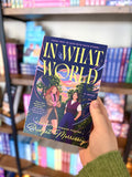 A hand holding a paperback copy of 'In What World' by Bridget Morrissey. The cover features a dark purple background with a colorful illustration of two women, and the title is written in large yellow letters.