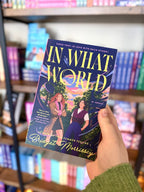 A hand holding a paperback copy of 'In What World' by Bridget Morrissey. The cover features a dark purple background with a colorful illustration of two women, and the title is written in large yellow letters.