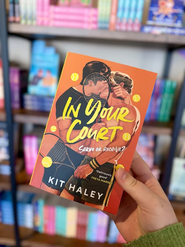 A hand holds a copy of the book 'In Your Court' by Kit Haley, featuring a bright orange cover with tennis-themed illustrations, and two men kissing.
