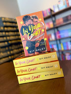 A stack of the book 'In Your Court' by Kit Haley, featuring a bright orange cover with tennis-themed illustrations, and two men kissing.
