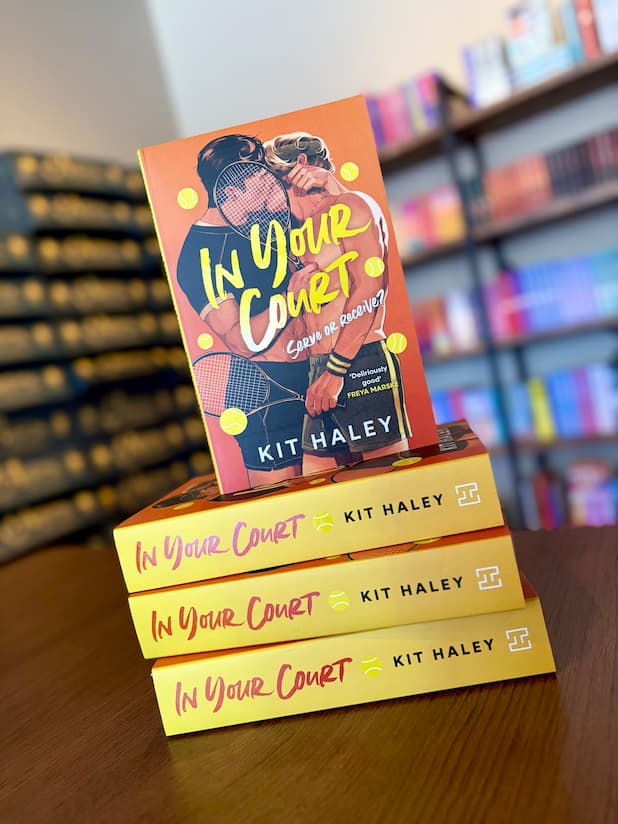 A stack of the book 'In Your Court' by Kit Haley, featuring a bright orange cover with tennis-themed illustrations, and two men kissing.
